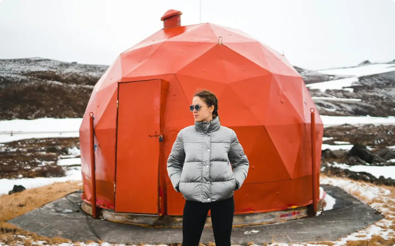 Persona con chaqueta de invierno de pie frente a una estructura geodésica roja en Islandia, simbolizando la aventura y la oportunidad de unirse al programa de afiliados de Cars Iceland.