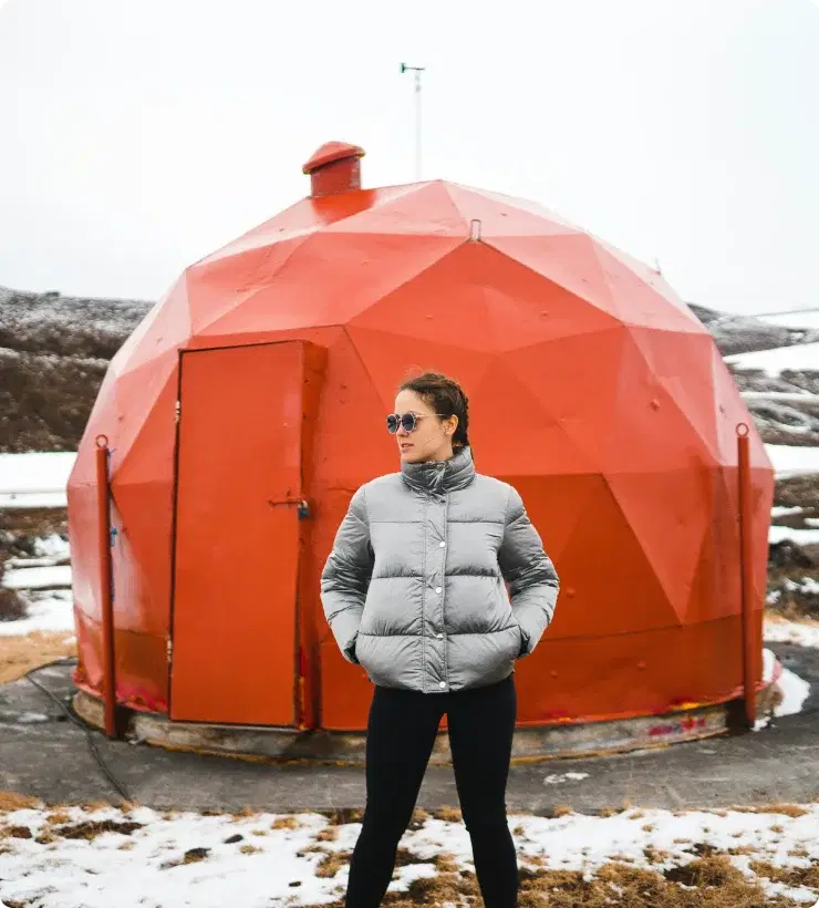 Persona con chaqueta de invierno de pie frente a una estructura geodésica roja en Islandia, simbolizando la aventura y la oportunidad de unirse al programa de afiliados de Cars Iceland.