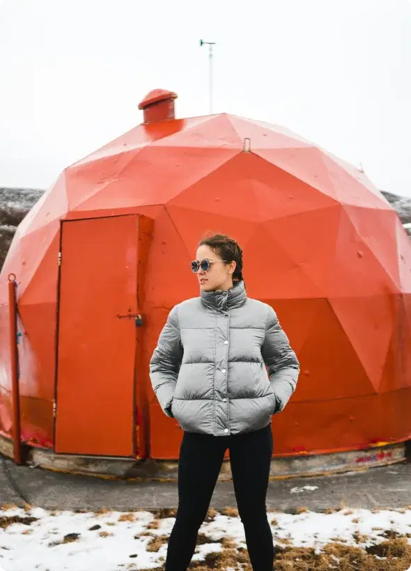 Persona con chaqueta de invierno de pie frente a una estructura geodésica roja en Islandia, simbolizando la aventura y la oportunidad de unirse al programa de afiliados de Cars Iceland.