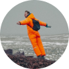Hombre con un equipo de invierno naranja brillante de pie sobre un terreno rocoso, con los brazos extendidos.