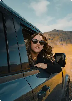 Mujer asomándose por la ventana de un auto con gafas de sol, disfrutando de un viaje panorámico a través del paisaje montañoso de Islandia.