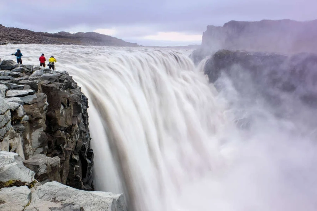 Observando la poderosa cascada Dettifoss en Islandia Tres personas con chaquetas coloridas están de pie al borde de un acantilado rocoso, observando la poderosa cascada Dettifoss en Islandia. La enorme cascada se precipita en el cañón nebuloso abajo, creando una escena dramática e impresionante. El cielo está nublado, añadiendo una atmósfera sombría al paisaje.