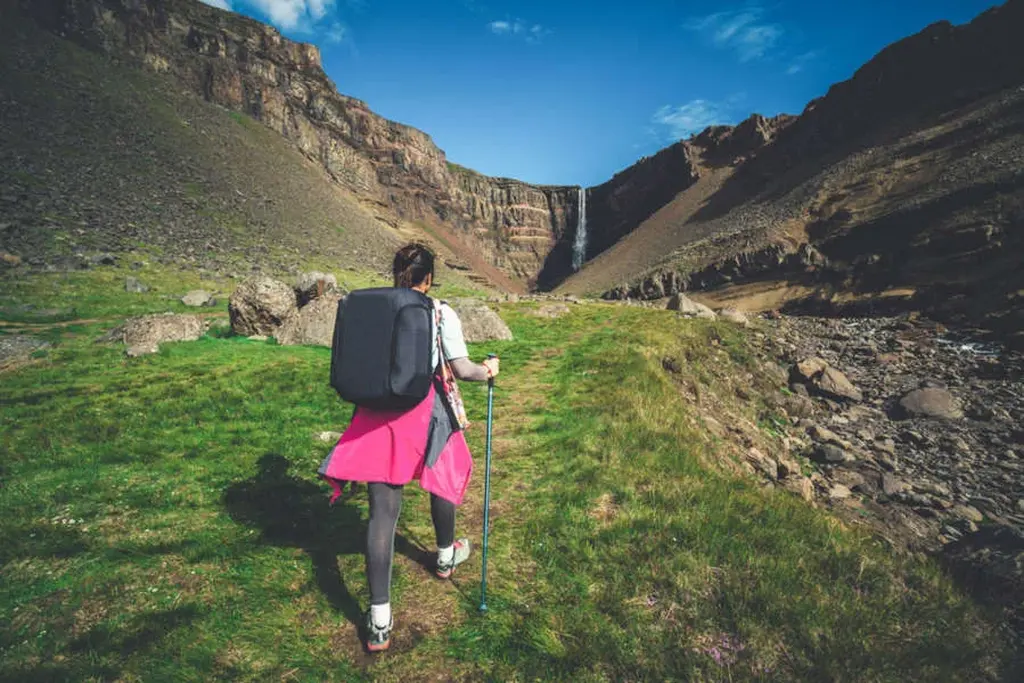 Caminata hacia la cascada Hengifoss en Islandia Un excursionista con una gran mochila y bastones de trekking caminando hacia la cascada Hengifoss en Islandia. El camino está rodeado de hierba verde exuberante, que contrasta con el terreno rocoso y las paredes del cañón que se elevan. La cascada cae por el acantilado a lo lejos, bajo un cielo azul brillante con algunas nubes dispersas.