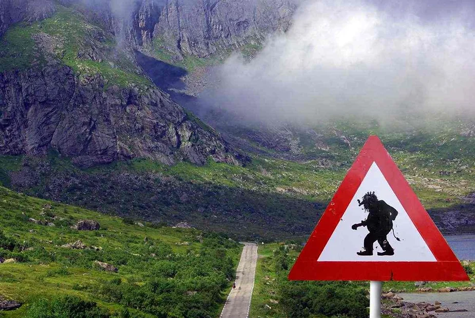 Una se&ntilde;al de tr&aacute;fico humor&iacute;stica que presenta la silueta de un troll y advierte sobre la presencia de trolls en la zona, con un tel&oacute;n de fondo de monta&ntilde;as brumosas y un camino sinuoso a trav&eacute;s de un terreno verde y exuberante.