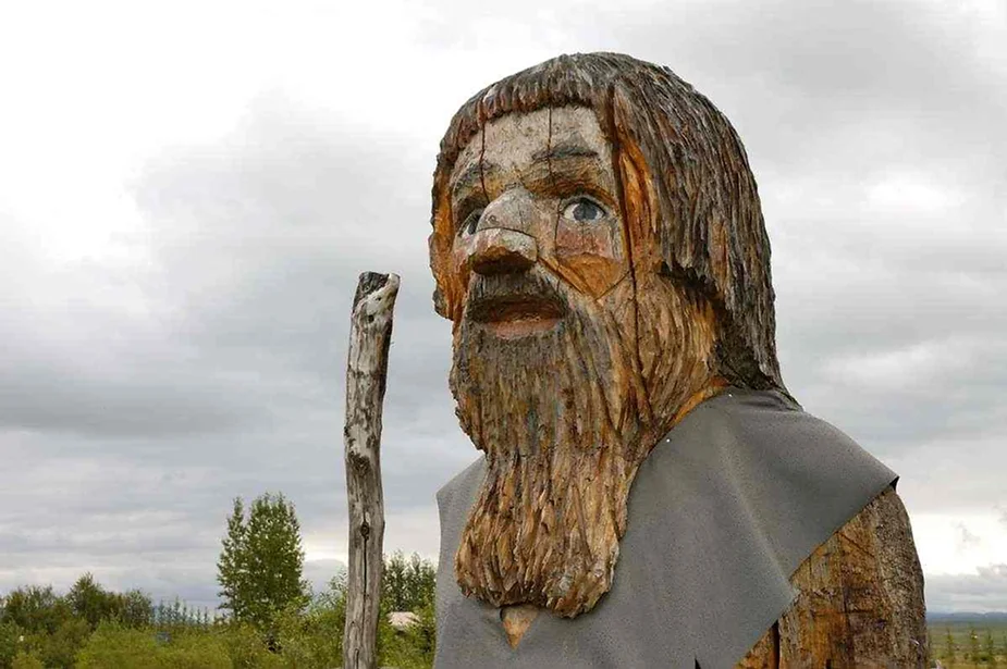 Un primer plano de una gran estatua de madera que representa a un hombre barbudo con rasgos r&uacute;sticos y tallados, de pie al aire libre bajo un cielo nublado, sosteniendo un bast&oacute;n de madera.