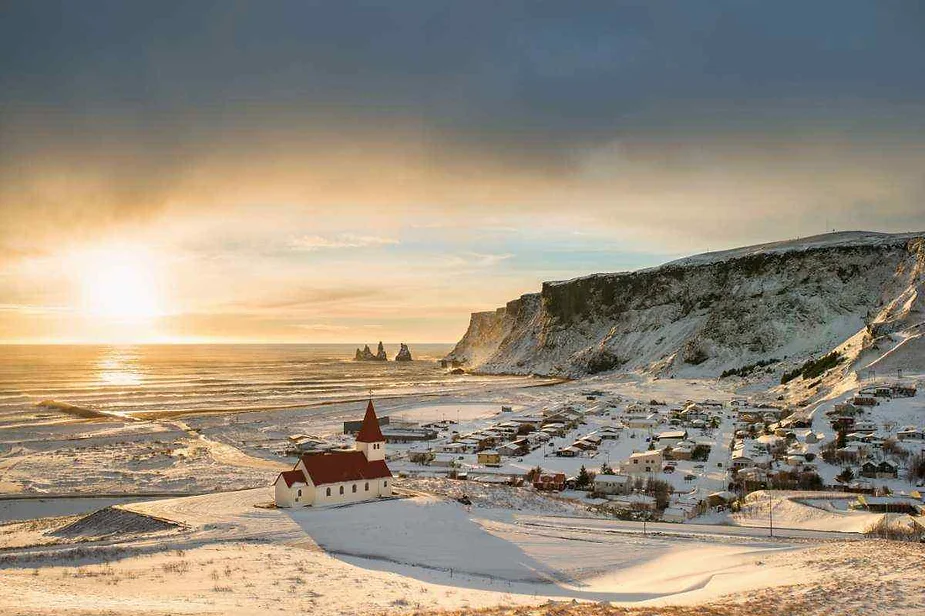Un pueblo costero cubierto de nieve en Islandia al amanecer, con una iglesia blanca con techo rojo en primer plano, acantilados espectaculares en el fondo y el sol proyectando un brillo dorado sobre el océano y el paisaje.