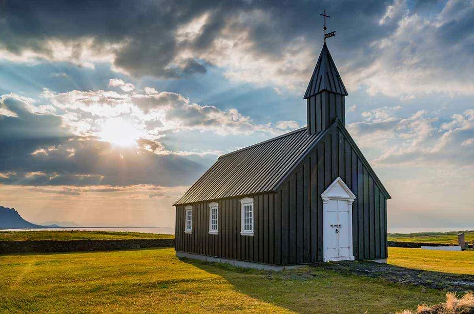 La iglesia de madera negra de Búðakirkja en Islandia, bañada por el cálido resplandor del sol poniente, con rayos que se abren paso a través de las nubes y un paisaje sereno de fondo.