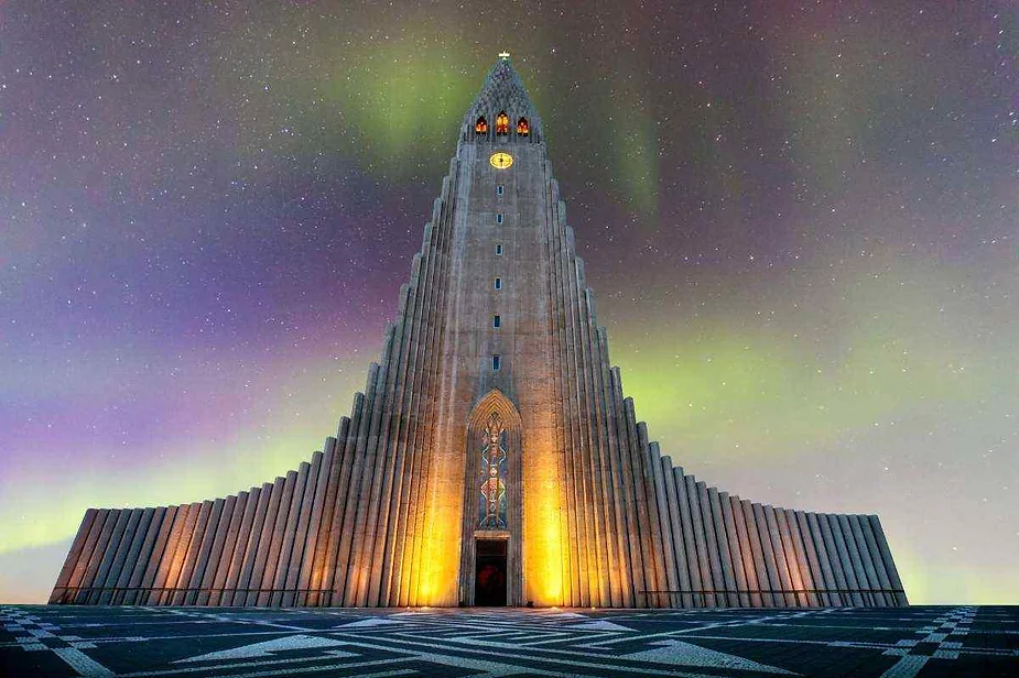 La fachada iluminada de la iglesia Hallgrímskirkja en Reykjavik, Islandia, bajo un cielo brillando con la aurora boreal, creando una escena nocturna impresionante y etérea.