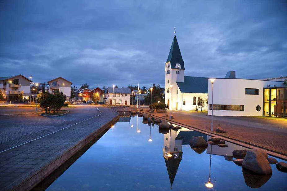 Una escena serena al atardecer en un pueblo islandés, con una iglesia blanca con techo verde reflejada en un estanque tranquilo, rodeada de calles y casas suavemente iluminadas bajo un cielo azul oscuro.