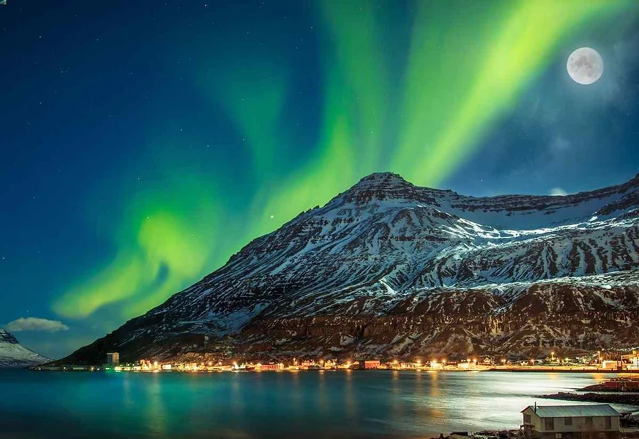 Auroras boreales iluminando el cielo nocturno sobre una monta&ntilde;a cubierta de nieve y un sereno pueblo island&eacute;s junto al agua, con la luna llena brillando intensamente.