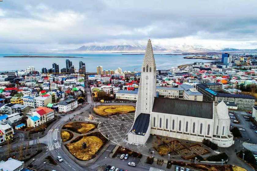 Vista a&eacute;rea de Reykjavik, Islandia, con la ic&oacute;nica iglesia Hallgr&iacute;mskirkja en primer plano, edificios coloridos y la costa con monta&ntilde;as al fondo bajo un cielo nublado.