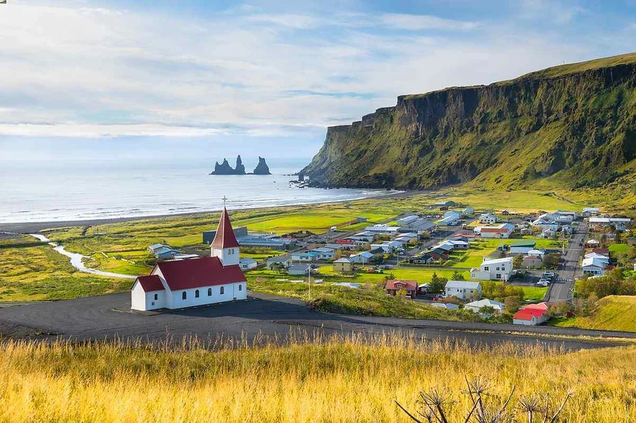 Vista panor&aacute;mica de un pueblo costero island&eacute;s con una iglesia con techo rojo, rodeada de exuberantes campos verdes y espectaculares acantilados con vistas al oc&eacute;ano.