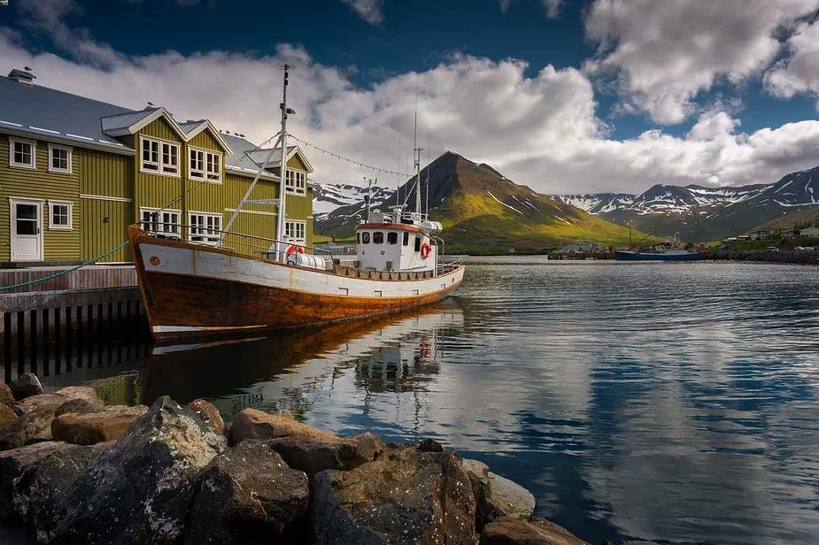 Una pintoresca escena portuaria en Islandia, con un barco de madera tradicional amarrado junto a edificios verdes, con monta&ntilde;as cubiertas de nieve reflejadas en el agua tranquila bajo un cielo espectacular.