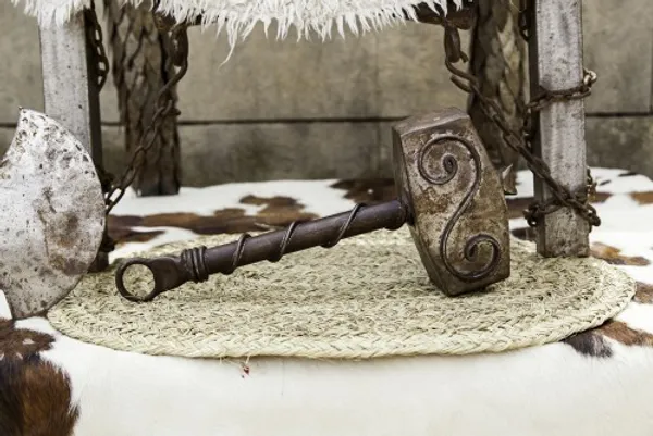 Martillo vikingo adornado con grabados en espiral en exhibición rústica Un gran martillo ornamentado que se asemeja al Mjölnir de Thor, elaborado en metal con intrincados patrones en espiral grabados en su cabeza, reposa sobre una estera tejida. El martillo está colocado cerca de una rústica silla de madera adornada con una manta de piel blanca. La escena también incluye un hacha de batalla desgastada que reposa cerca, evocando un tema vikingo o nórdico.