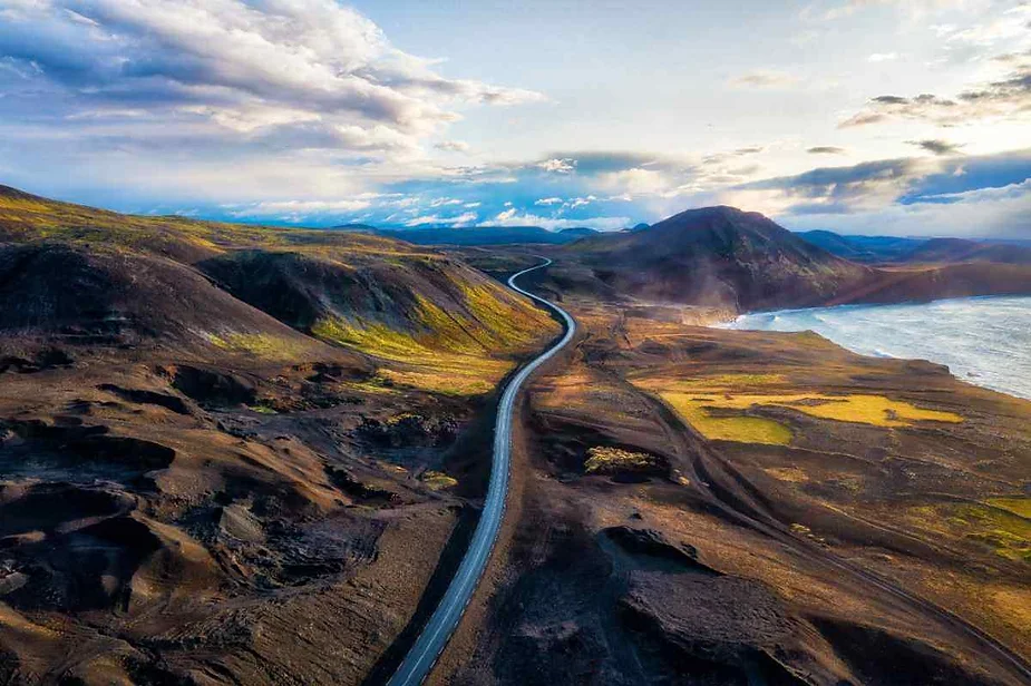 Una carretera serpenteante se extiende por el escarpado paisaje volcánico de Islandia, con colinas estériles y una vista lejana de la costa bajo un cielo dramático.