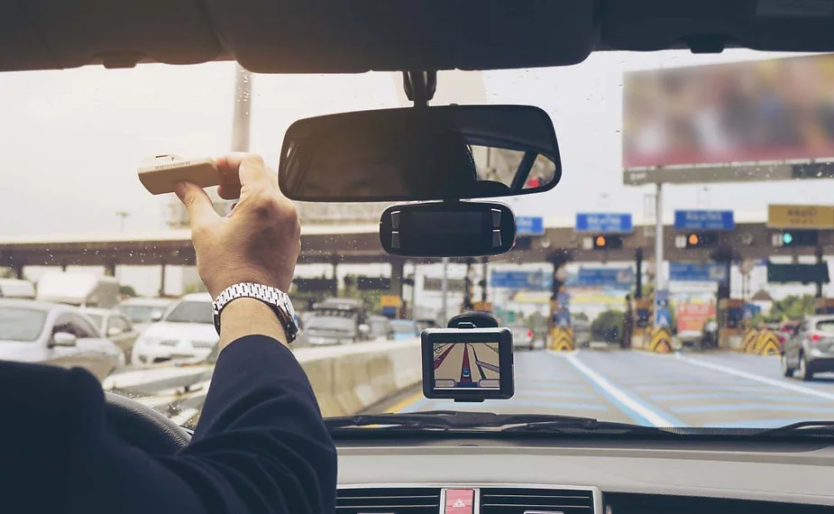 Un conductor dentro de un coche acerca la mano al parabrisas con un dispositivo de peaje, preparándose para entrar en un carril de cabina de peaje; en el salpicadero, un GPS muestra la ruta y se ven otros vehículos delante.