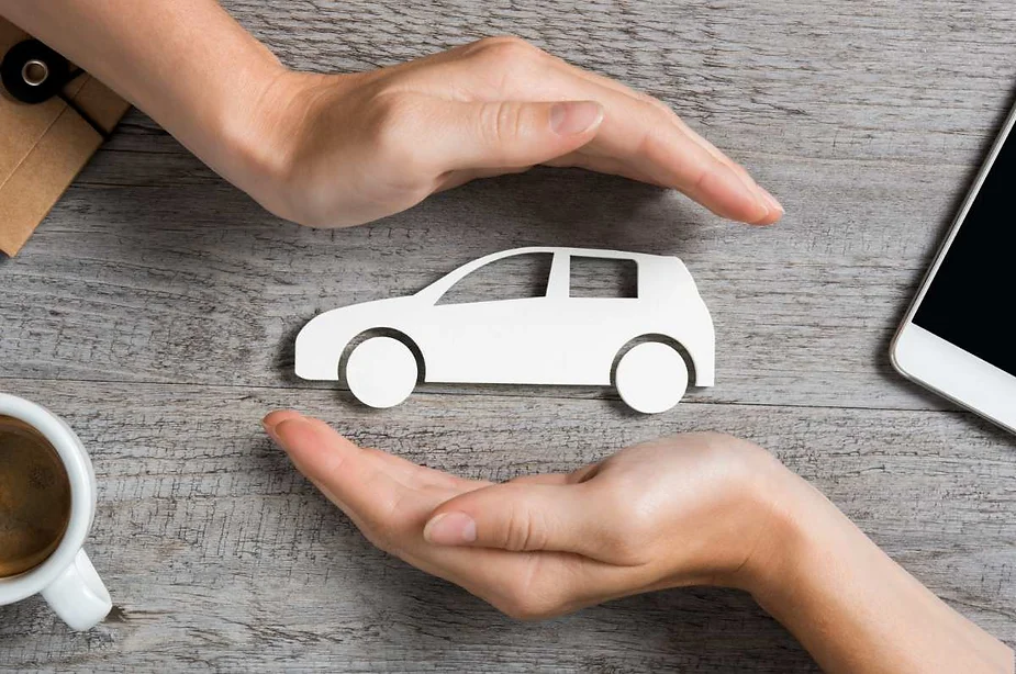 Manos protegiendo un pequeño recorte de papel de un coche sobre una superficie de madera, simbolizando el seguro de coche y la protección del vehículo
