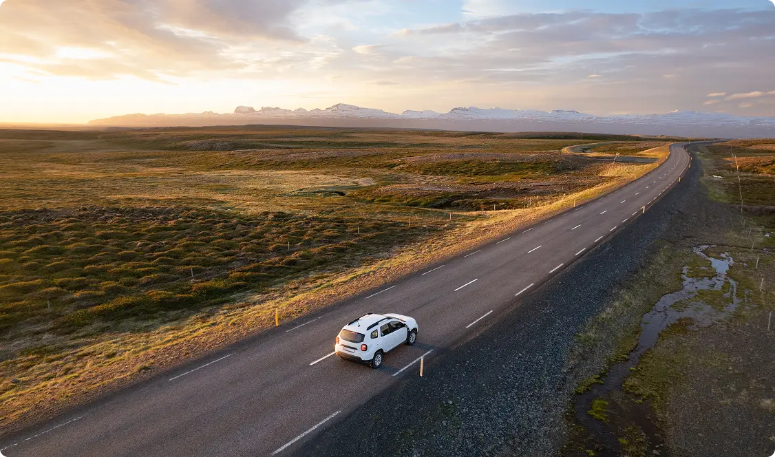 Vista aérea de un río serpenteante y las agrestes tierras altas de Islandia, con un coche cruzando un puente, simbolizando la aventura y la libertad de explorar Islandia con un coche de alquiler.